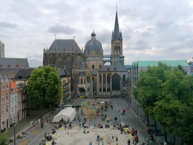 View over Aachen Cathedral and the surrounding square with historic buildings, trees, and people gathering in the city center.